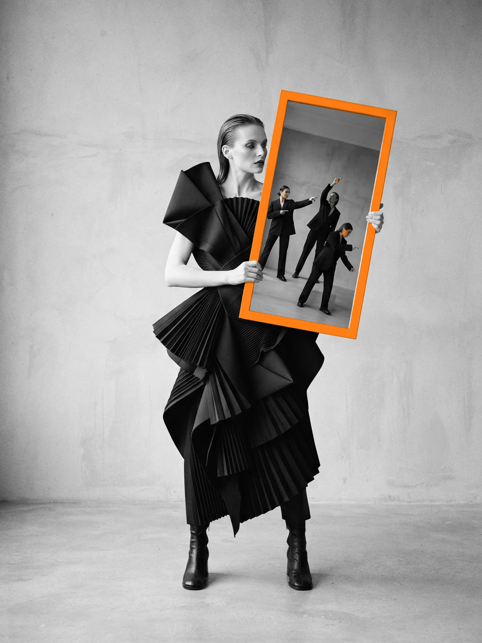 A woman in a sculptural black paper dress holds an orange-framed mirror that reflects three people pointing in different directions.