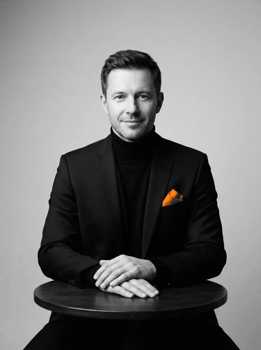 A man in a dark jacket sits at a small round table, hands folded, an orange pocket square in his jacket.